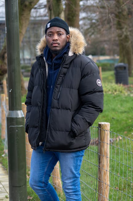 A young man with dark skin is standing outdoors in a park-like setting, dressed in a thick black puffer jacket with a fur-lined hood, a navy blue hoodie underneath, and a black knit beanie on his head. His hands are in the jacket pockets, and he has a serious expression while looking at the camera. In the background, there are green grassy areas, a wooden fence with wire mesh, and trees with bare branches suggesting late autumn or winter. To the left, a metal pole with a metallic cap is visible, and further back, there are more trees and a few indistinct structures, possibly park furniture or bins. The environment appears clean and well-maintained, with natural lighting illuminating the scene evenly. This image depicts an individual dressed warmly, potentially in a setting where outdoor activity or casual walks require suitable attire, and reflects a typical urban outdoor environment possibly associated with waste disposal or community spaces, in line with the services of Waste Disposal Croydon.