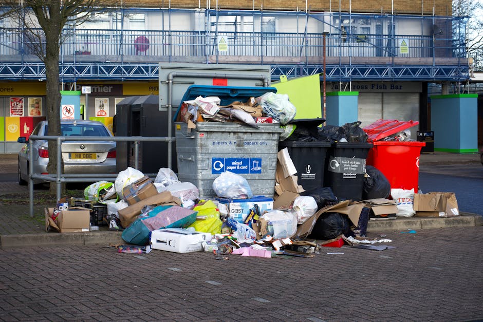 A large collection of overflowing rubbish and waste materials piled beside a commercial waste disposal bin, located on a paved urban street. The waste includes crumpled cardboard boxes, plastic bags, paper packaging, and various discarded packaging materials, some spilling onto the surrounding pavement. The bin is made of metal with a blue lid and has printed labels indicating it is for mixed paper and card. Nearby, there are black and red waste bags and additional cardboard boxes, some flattened and others partially open. In the background, there is a blue scaffolding structure covering the facade of a building, and storefronts with closed shutters, signifying a commercial area. A parked silver car is visible on the left side, and a tree with a bare trunk stands to the left. The scene is lit by natural daylight, highlighting the contrast between the clean street environment and the unmanaged waste, exemplifying the importance of proper rubbish collection and disposal services, such as those provided by Waste Disposal Croydon, to prevent illegal dumping or cluttering in urban areas.