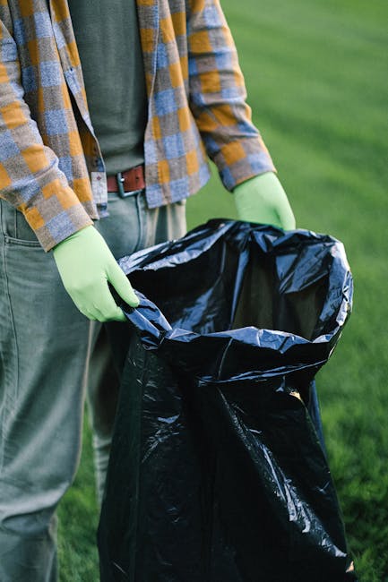 A young man with dark skin is standing outdoors in a park-like setting, dressed in a thick black puffer jacket with a fur-lined hood, a navy blue hoodie underneath, and a black knit beanie on his head. His hands are in the jacket pockets, and he has a serious expression while looking at the camera. In the background, there are green grassy areas, a wooden fence with wire mesh, and trees with bare branches suggesting late autumn or winter. To the left, a metal pole with a metallic cap is visible, and further back, there are more trees and a few indistinct structures, possibly park furniture or bins. The environment appears clean and well-maintained, with natural lighting illuminating the scene evenly. This image depicts an individual dressed warmly, potentially in a setting where outdoor activity or casual walks require suitable attire, and reflects a typical urban outdoor environment possibly associated with waste disposal or community spaces, in line with the services of Waste Disposal Croydon.