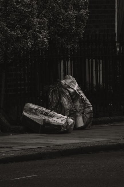 Three large black plastic rubbish bags filled with waste are placed on the pavement next to a black metal fence, with a small tree or shrubbery visible behind. The bags appear to be tightly packed and slightly crumpled, indicating they are ready for collection or disposal. The scene is set outdoors on a street area, with a smooth asphalt surface in the foreground and a curb separating the pavement from the roadway. The lighting suggests overcast or low-light conditions, contributing to the overall muted tone of the image. The image relates to private waste handling and rubbish removal services, such as those offered by Waste Disposal Croydon, providing visual context for local excess waste clearance in an urban environment.