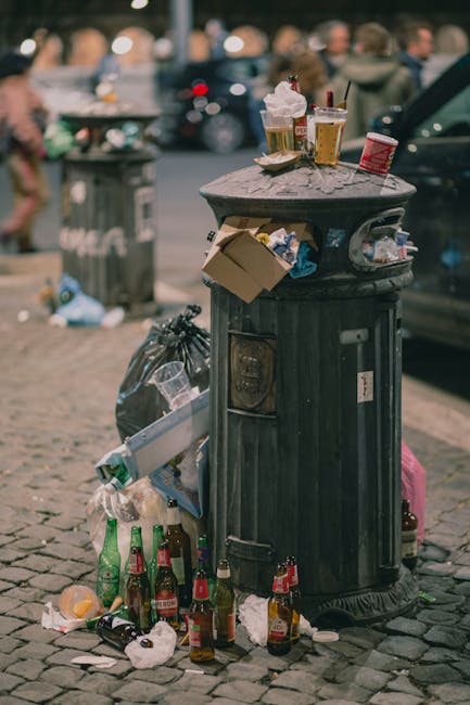 Three large black plastic rubbish bags filled with waste are placed on the pavement next to a black metal fence, with a small tree or shrubbery visible behind. The bags appear to be tightly packed and slightly crumpled, indicating they are ready for collection or disposal. The scene is set outdoors on a street area, with a smooth asphalt surface in the foreground and a curb separating the pavement from the roadway. The lighting suggests overcast or low-light conditions, contributing to the overall muted tone of the image. The image relates to private waste handling and rubbish removal services, such as those offered by Waste Disposal Croydon, providing visual context for local excess waste clearance in an urban environment.