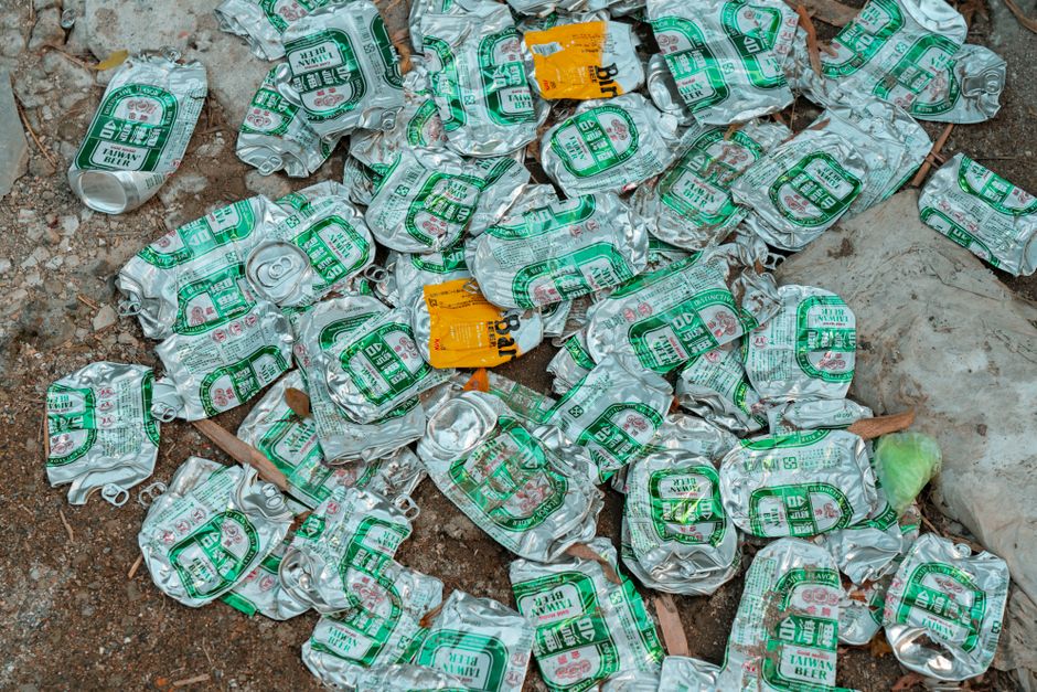 A pile of crushed aluminum cans with a silver, metallic finish, featuring green and white printed labels, scattered on a dirt ground with small rocks and dry leaves. Some cans are partially folded or bent, showing their tapered openings. In the background, there are a few pieces of trash, including a yellow paper label and a plastic bag. The scene appears to be outdoors, possibly on a sidewalk or garden area, where waste has been discarded or collected for disposal by a waste management service like Waste Disposal Croydon. The overall environment is informal, with the cans loosely piled, reflecting typical discarded rubbish that may be targeted for removal or recycling through independent waste collection or private waste disposal solutions. The focus is on the metallic, reflective surfaces of the cans and the natural earthy tones of the ground and surrounding debris, emphasizing a scene of outdoor waste accumulation.