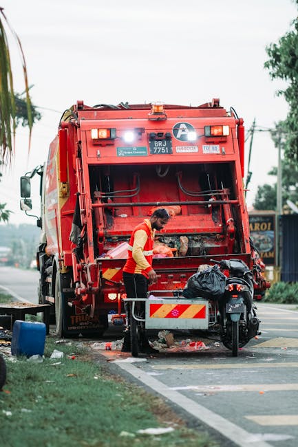 A waste disposal worker wearing a high-visibility yellow and orange vest is positioned behind a large red rubbish collection truck on a street, with the back panel open, revealing the internal compaction mechanism. The worker is holding a black trash bag, which is being fed into the vehicle, while standing beside a small metal stand with a reflective striped warning sign. In the foreground, there is a black motorbike parked on the pavement, partially obstructing the view of the worker's lower body. The street scene includes a curb, a patch of grass, and a blue plastic container on the side of the road. In the background, a row of trees and utility poles are visible, with overhead power lines crossing the sky, which is partly cloudy. The image demonstrates typical street-level waste collection activity related to private rubbish removal services, reflecting an on-site or street-side approach to waste disposal consistent with independent collection methods often used by companies like Waste Disposal Croydon.