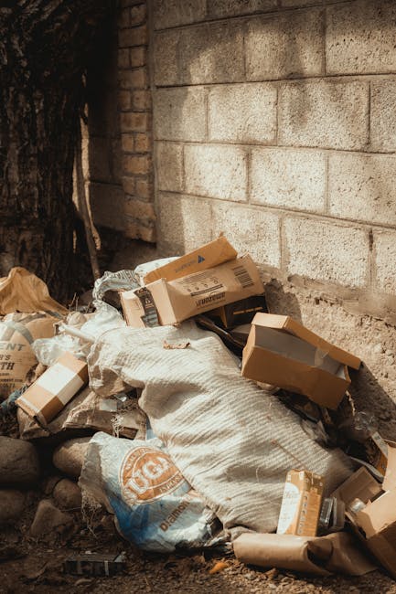 A large pile of mixed household rubbish and waste materials accumulated around three bins in a parking area adjacent to a commercial building. The debris includes cardboard boxes, paper bags, plastic packaging, and black and red refuse bags, with some items spilling onto the paved ground. The central bin, made of metal with a sliding lid, is partially open and overflowing with discarded paper, cardboard, and plastic waste. To its right are two black bins and a red bin, all filled with rubbish and tightly closed. Behind the rubbish, a metal railing separates the area from a parked silver car with a yellow registration plate, positioned on a paved driveway. In the background, the building's facade features shopfronts with closed shutters, storefront signage, and a partly visible blue scaffolding structure. The scene is lit by natural daylight, illustrating an example of unmanaged waste accumulation in an urban environment that may require professional waste disposal services such as those provided by Waste Disposal Croydon to manage rubbish effectively.