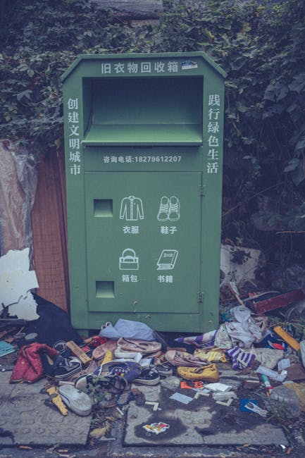 A green, metal clothes collection box with Chinese characters at the top, positioned on a sidewalk in front of a natural, wooded background. The box has a rectangular opening near the top for deposit and additional compartments below for specific items such as clothing, shoes, bags, and books, each labeled with icons and Chinese text. The surface of the deposit box appears slightly weathered, with some minor rust and paint chipping visible around the edges. Surrounding the base of the box, there is a significant amount of scattered rubbish including clothing, shoes, plastic packaging, and other miscellaneous waste materials, creating a cluttered scene on the pavement. The environment suggests an outdoor location with natural foliage in the background, and the pile of rubbish indicates improper disposal or an overflow of waste collection, highlighting a challenge for local waste management services like Waste Disposal Croydon, which may be involved in alternative waste handling or rubbish removal efforts in the area.