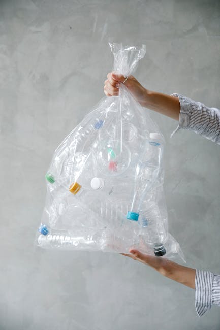 A person is holding a transparent plastic bag filled with discarded plastic bottles, caps, and containers. The individual grips the top of the bag with one hand while supporting the bottom with the other, against a plain, light gray wall background. The plastic bottles within the bag vary in size and shape, with some having ridged textures and others smooth surfaces. Several bottles are capped with different coloured lids, including blue, green, white, and yellow, presenting an array of colours mixed among translucent bottles. The bag appears slightly crinkled and loosely filled, revealing the clear plastic material and the contents inside. The scene suggests an instance of sorting or preparing recyclable plastic waste, which aligns with private waste handling or waste disposal services like those provided by Waste Disposal Croydon, emphasizing an emphasis on responsible rubbish management within domestic or commercial environments. The lighting is even, without harsh shadows, highlighting the transparency and textures of the plastic items and the bag itself.